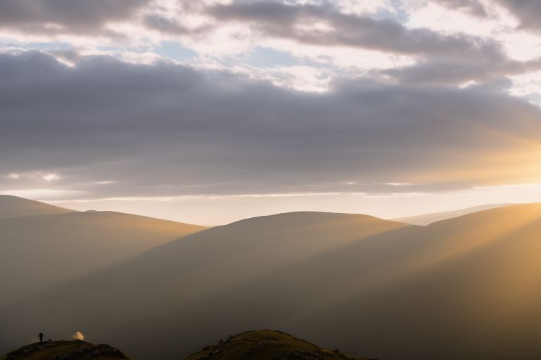 A person standing atop a mountain overlooking rolling hills with rays of sunshine peeking through clouds above them