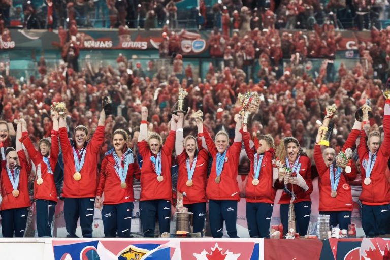 A picture of the winning Canadian team celebrating with medals around their necks while holding up trophies above their heads, surrounded by cheering fans in a stadium setting