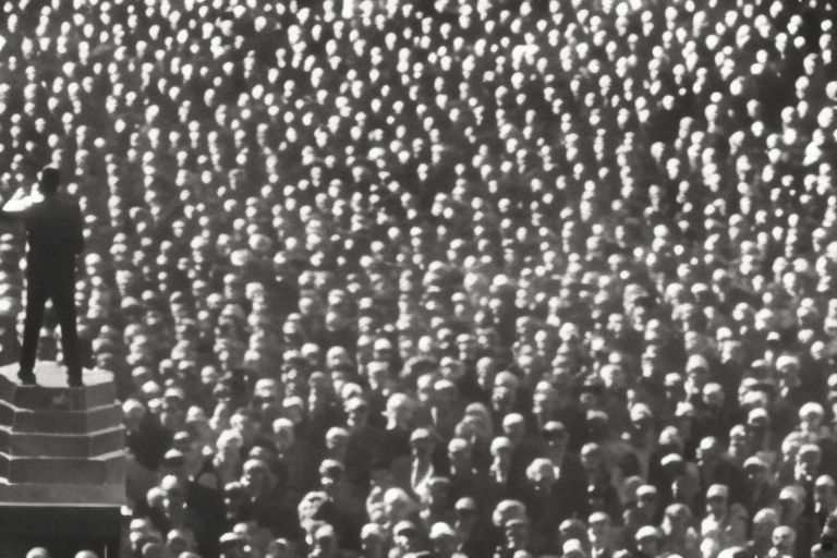 A silhouette of an individual standing alone on a podium speaking into a microphone with hundreds of people around him looking up.