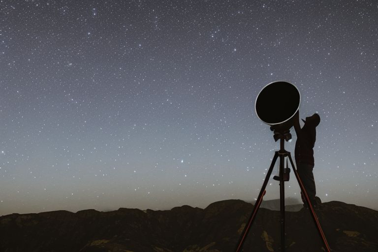 An image of a person looking through a telescope at night sky with stars twinkling above them