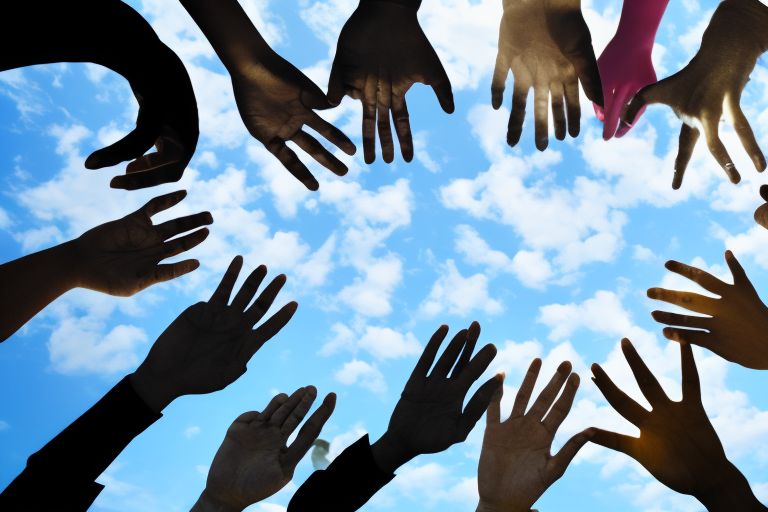 An image showing several hands raised against a bright blue sky with clouds forming into the shape of a heart around them representing unity amongst diversity