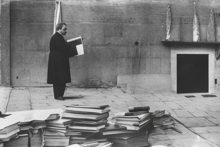Immagine di Luigi Rigolio mentre parla a gruppodi personecircondate da varioggetti simbolicicome un libro antico ,una matita ,una melaa metà tagliata ecc..